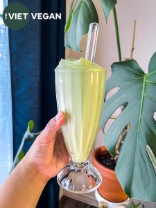 avocado smoothie in a milkshake glass with a glass straw, held in front of some houseplants