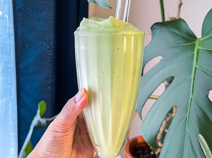 avocado smoothie in a milkshake glass with a glass straw, held in front of some houseplants