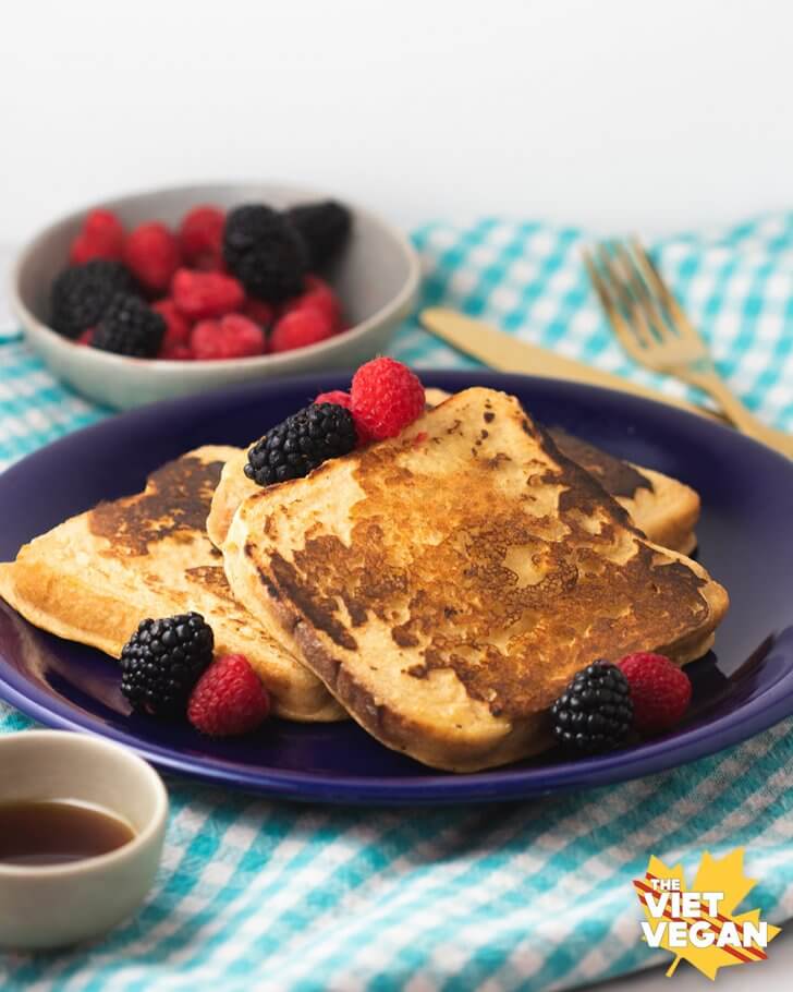 Vegan French toast with berries, maple syrup in a little cup and gold cutlery on the side.