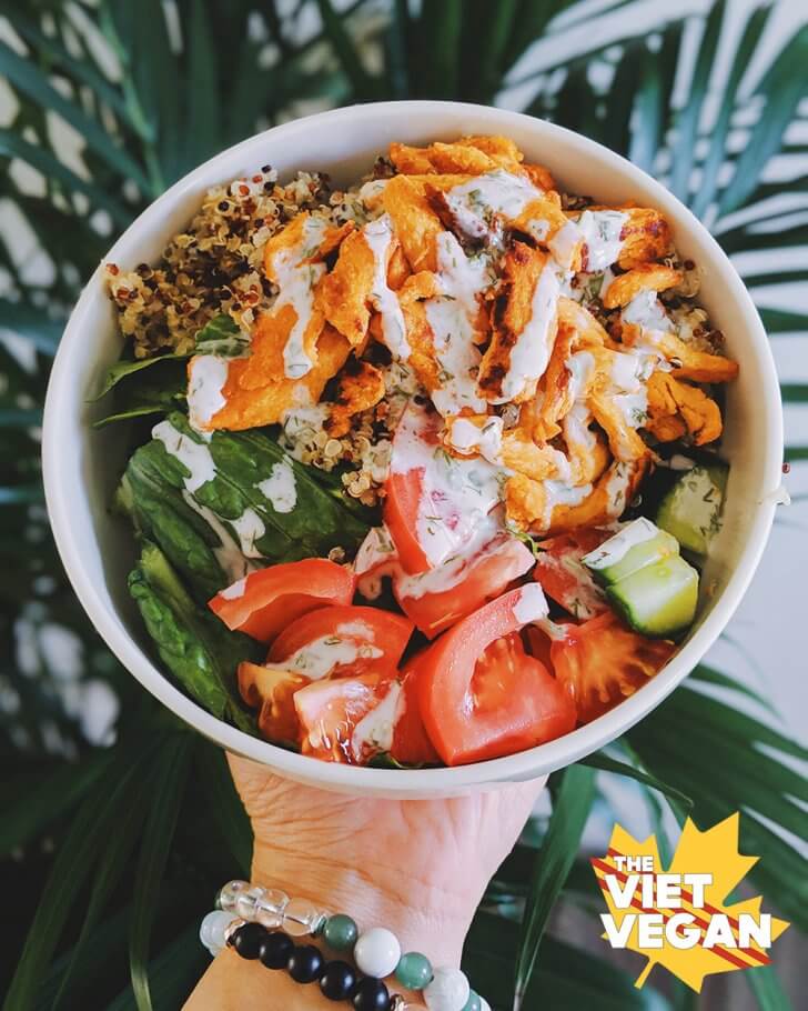 Vegan buffalo chicken in a bowl with quinoa, tomato, cucumber, and romaine lettuce. Palm in the background.