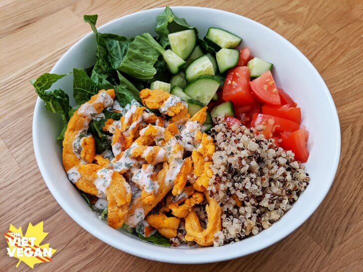 Vegan buffalo chicken in a bowl with quinoa, tomato, cucumber, and romaine lettuce. Bowl on wood.