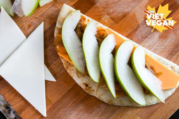 A top view of sliced pear layered on top of caramelized onions, vegan cheese, and half moon of naan bread on a cutting board