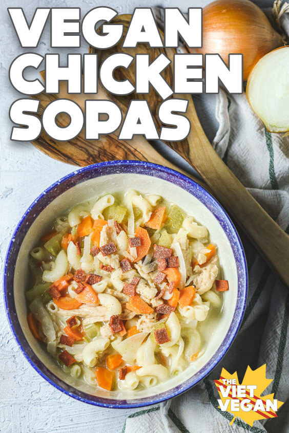 Vegan chicken sopas (macaroni soup) in a blue rimmed bowl with wooden ladle and an onion around it