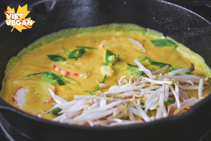 vegan banh xeo Vietnamese savoury pancake in pan cooking, with vegan shrimp and onions embedded in the batter, mung bean sprounts on one half