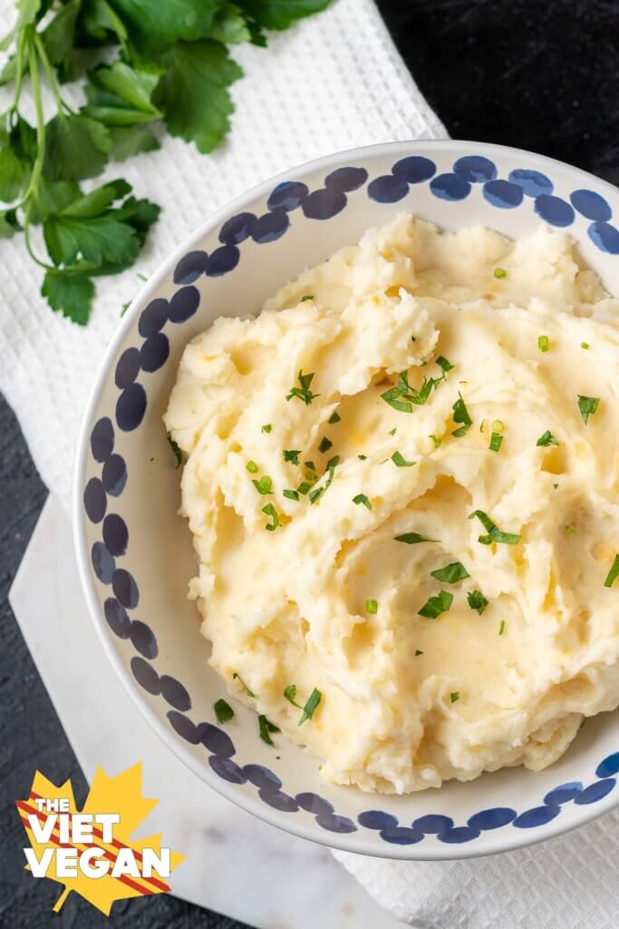 Super fluffy vegan mashed potatoes in a while bowl with blue accents around the rim, with parsley on top and on the side of the photo