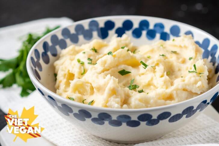 Horizontal shot of super fluffy vegan mashed potatoes in a while bowl with blue accents around the rim, with parsley on top and on the side of the photo