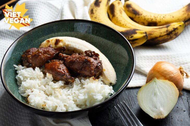 vegan adobo drumsticks in a bowl with rice and a banana, a sliced onion and whole bananas around for decoration