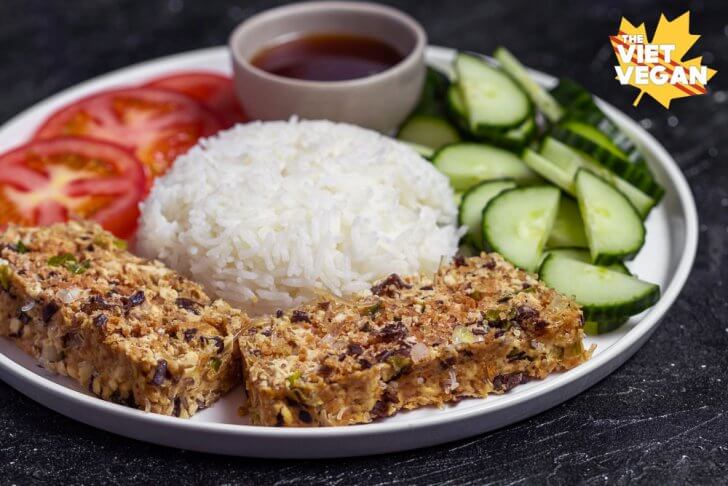 meatloaf slices on a plate with a mound of white rice, cucumber, sliced tomato, and sauce, shot from the side