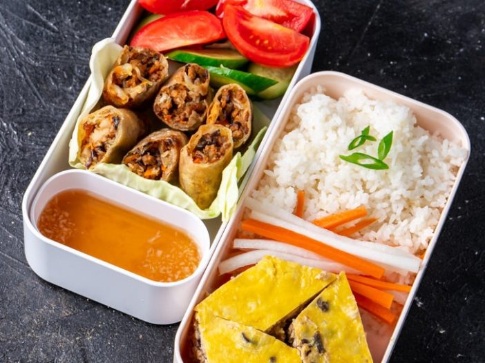 Vietnamese bento with two layers, one with rice, egg meatloaf, pickled carrots and daikon, the other with spring rolls and vegetables and sauce