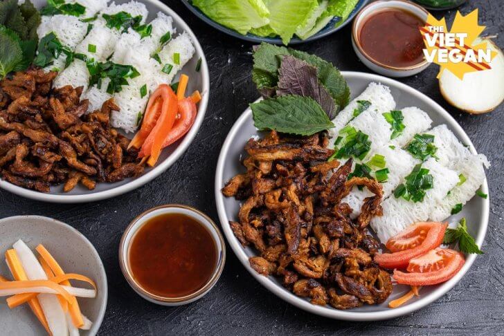 Rice vermicelli noodle bundles on a plate with panfried soy curls, tomato, red mint, with sauce, lettuce, and pickled carrots & daikon in the background, horizontal shot on a black background