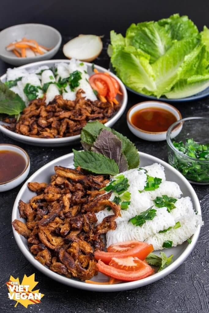 Rice vermicelli noodle bundles on a plate with panfried soy curls, tomato, red mint, with sauce, lettuce, and pickled carrots & daikon in the background