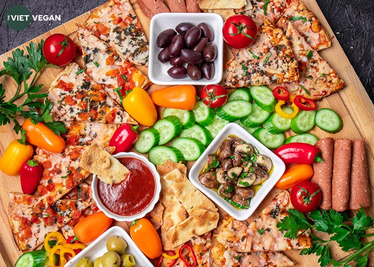 Overhead shot of snack board, three bowls contain black olives, green olives, sauce, and marinated mushrooms, and are surrounded by baked Wholly Veggie Pizza and sliced fresh veggies