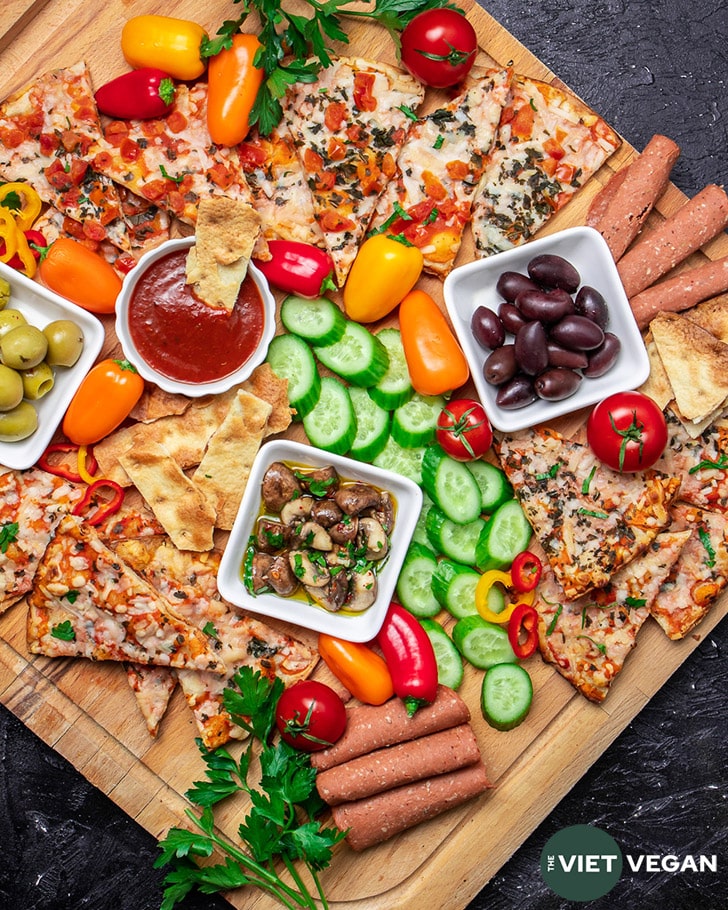 Overhead shot of snack board, three bowls contain black olives, green olives, sauce, and marinated mushrooms, and are surrounded by baked Wholly Veggie Pizza and sliced fresh veggies