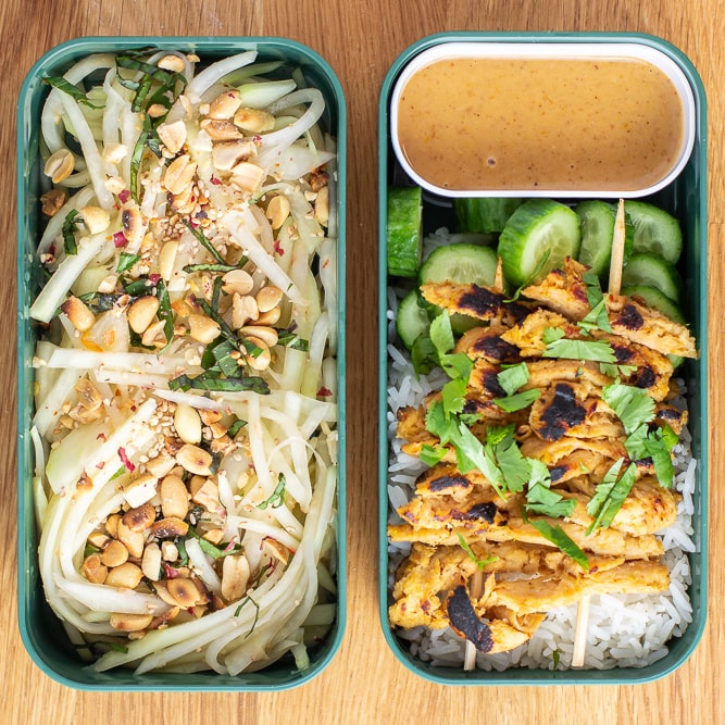 Two rectangular bento containers with green papaya salad in the left, and rice, peanut sauce, cucumbers, and satay soy curl skewers on the right