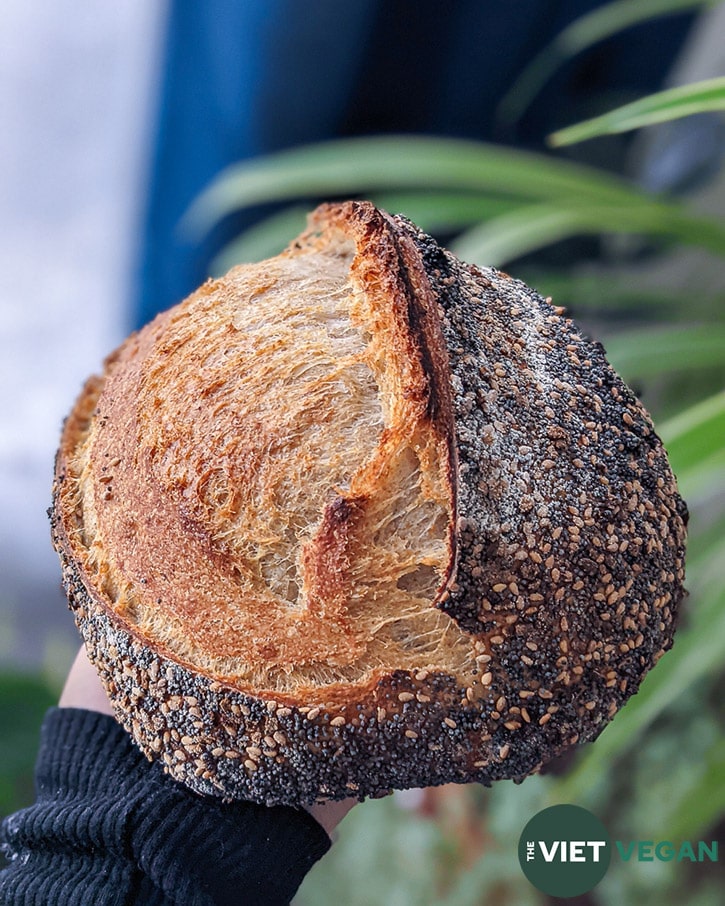 A hand holding up a sourdough loaf that has poppyseeds, dried onion flakes, and sesame seeds on the outer crust