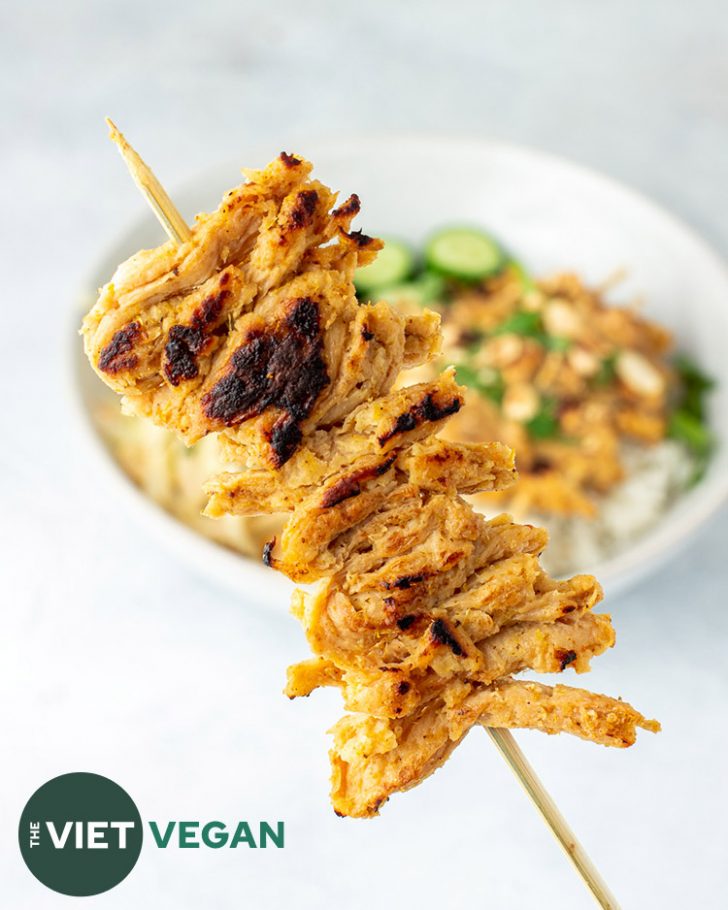 satay on a stick in foreground, with bowl of satay, rice, and green papaya salad in the background