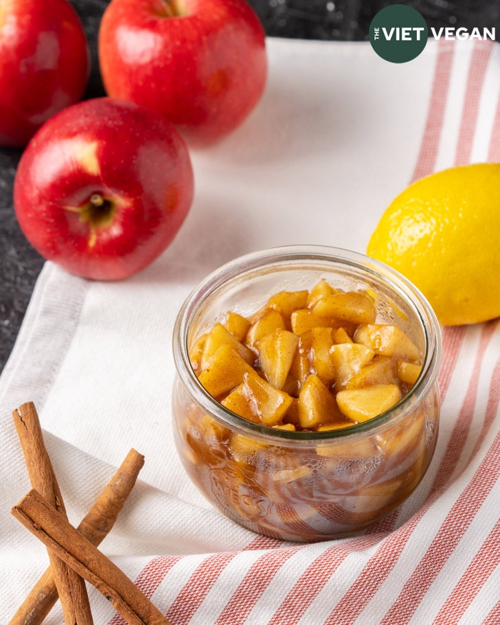 apple pie filling in jar surrounded by apples, lemon, and cinnamon