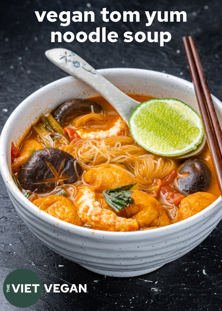 a bowl of tom yum noodle soup with a lime wedge resting on a spoon to squeeze on top, with wooden chopsticks on the edge of the bowl.