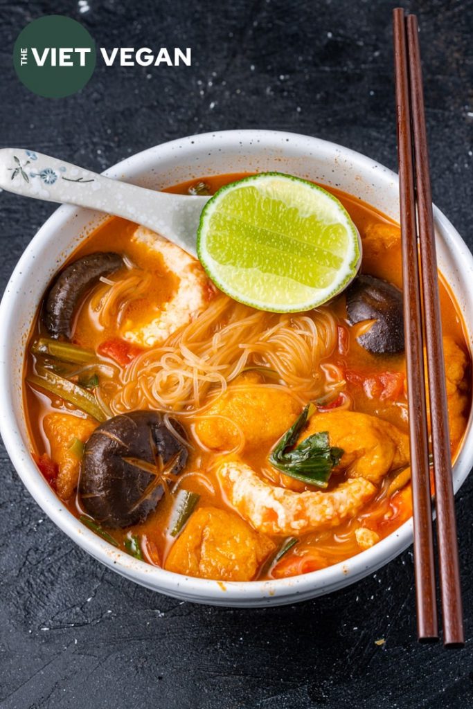 overhead photo of tom yum noodle soup with lime, mushrooms, vegan shrimp, noodles, tomatoes, and wooden chopsticks on the edge of the bowl