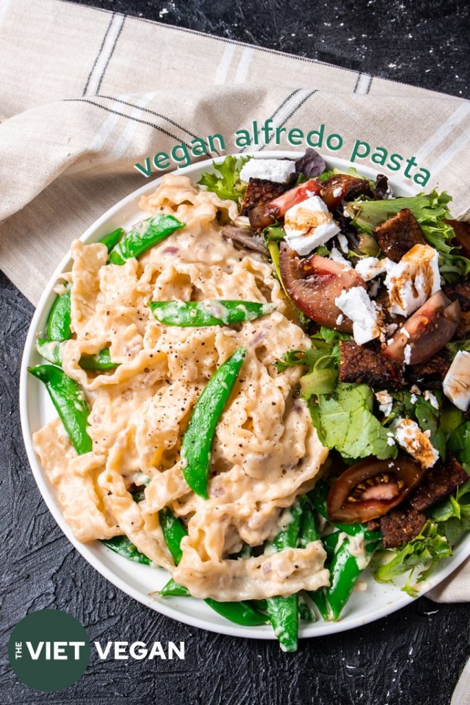 vegan alfredo pasta on a plate with salad with a beige plaid tea towel on a dark background, vertical angle