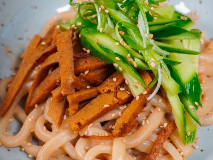 sesame udon topped with sliced seitan, cucumber, and green onion curls