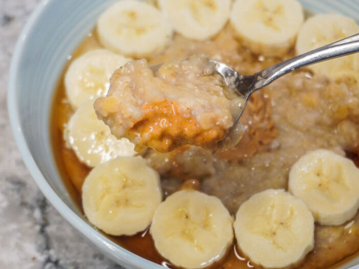 a blue bowl filled with red lentil oats, topped with banana and maple syrup. A spoonful of oatmeal with peanut butter over the bowl.